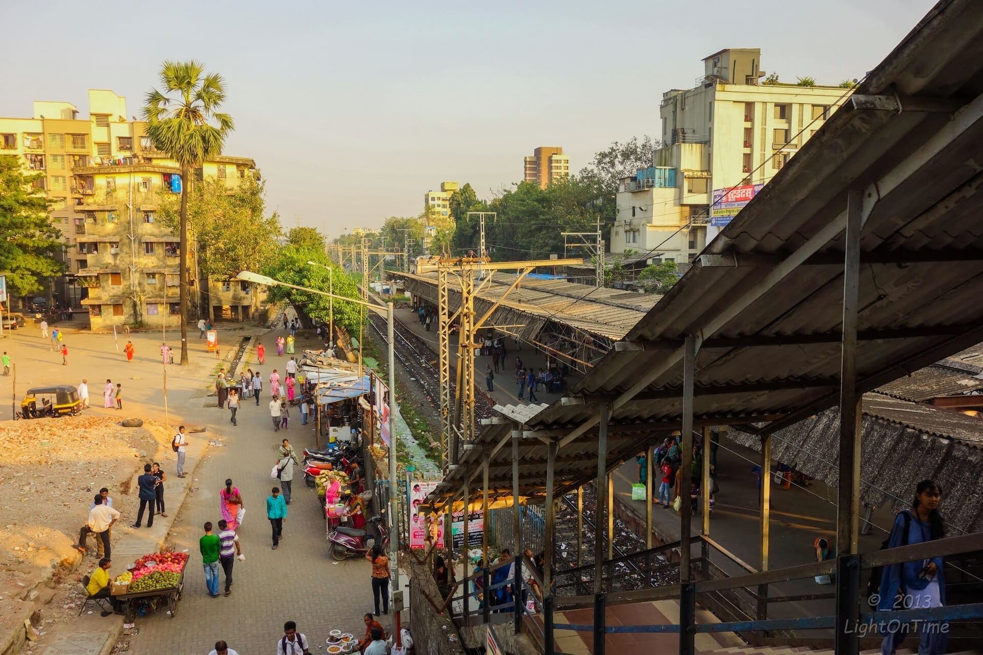 Chembur station seen from the footbridge, with a market street beside the platform roof in evening light