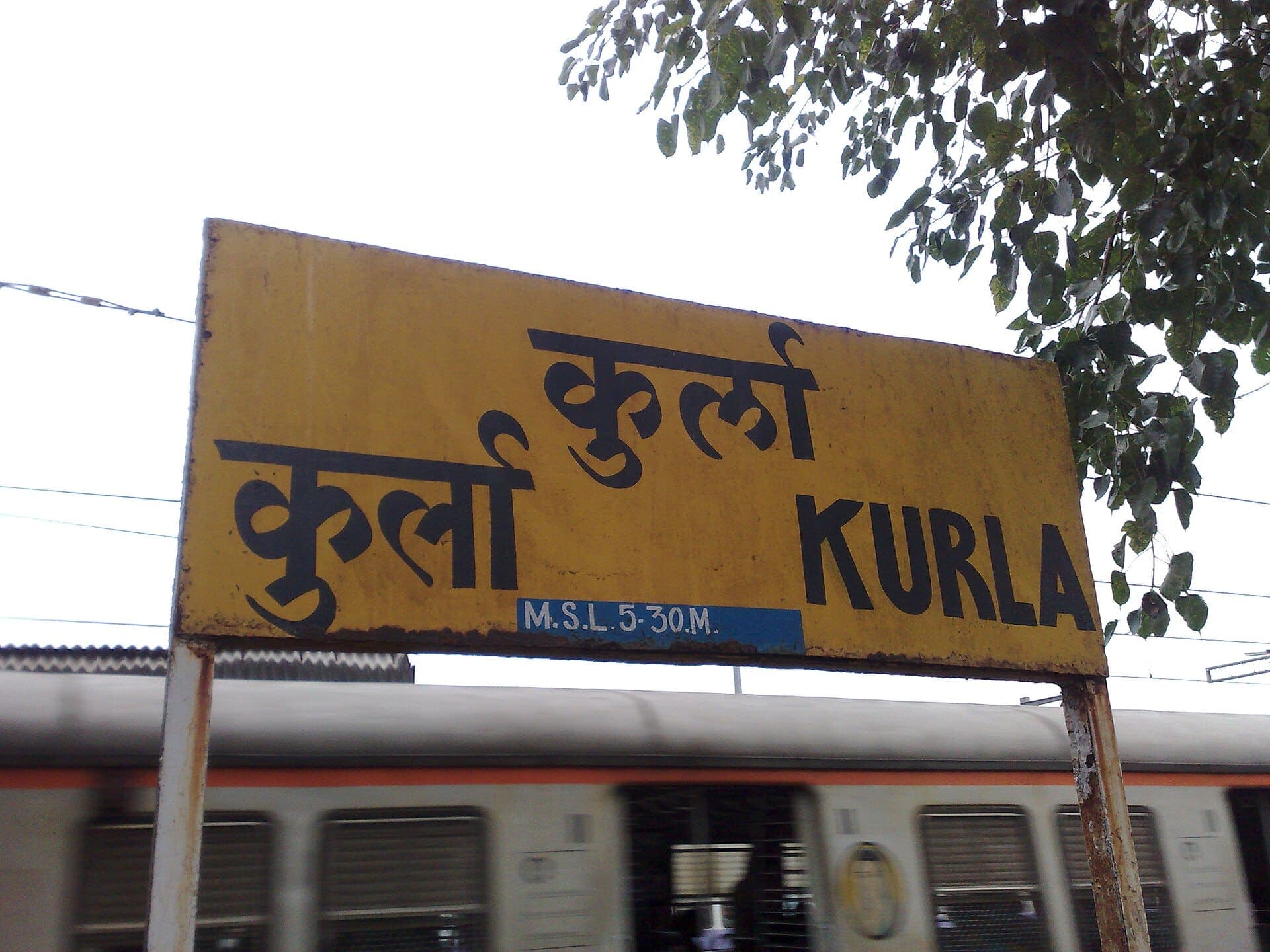 The yellow name board of Kurla station in Marathi, Hindi and English, with a local train behind it