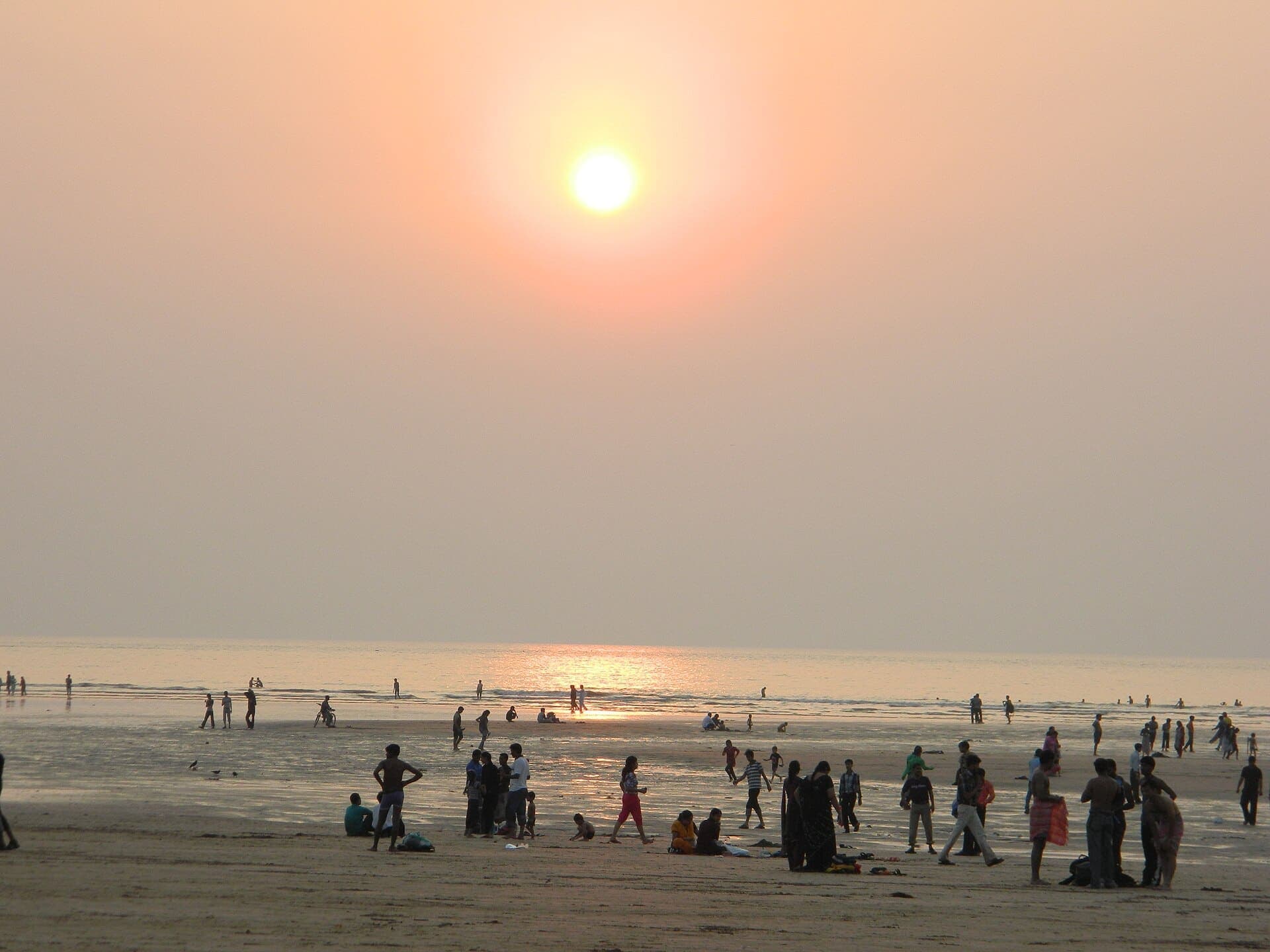People walking on the wet sand of Aksa Beach under a low orange sun